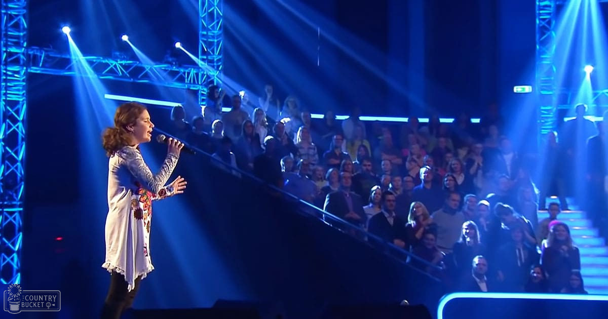 An adult male singer performing on a tiered indoor stage in front of an engaged audience with professional lighting setups.