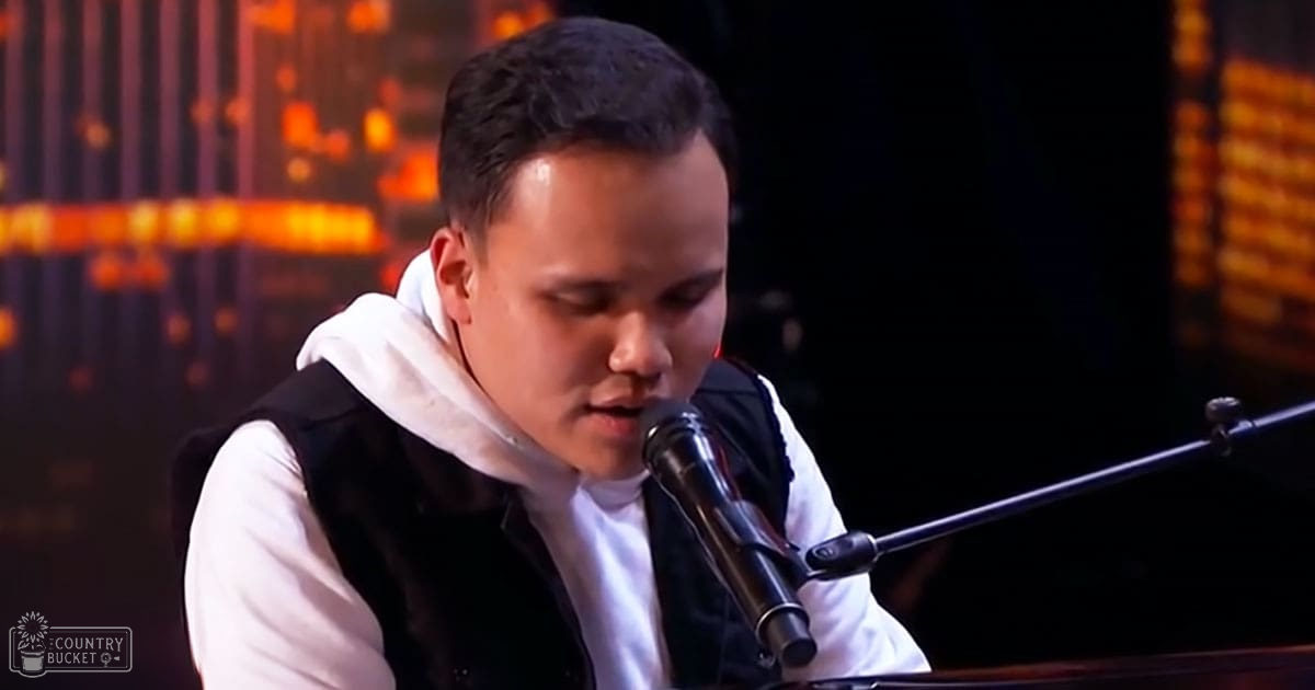 A young adult male performer wearing a white hoodie with black sleeves and collar, standing at an indoor event with stage lighting.