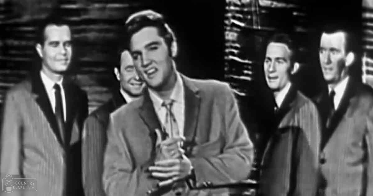 A young adult male singer at center, wearing a tailored suit and tie, surrounded by other similarly dressed individuals in an indoor performance setting.