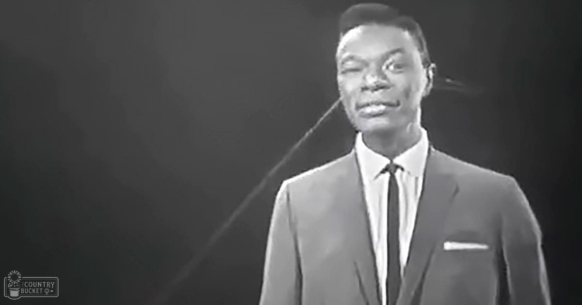 A young adult male wearing a mid-20th century suit with a pocket square stands in an indoor setting, possibly on stage or for a performance.