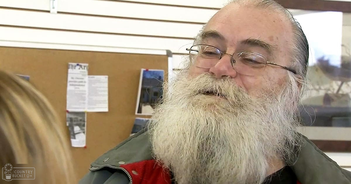 An older adult male with a long white beard and glasses, wearing a dark green jacket over a red garment inside what seems to be a community center or similar indoor setting.