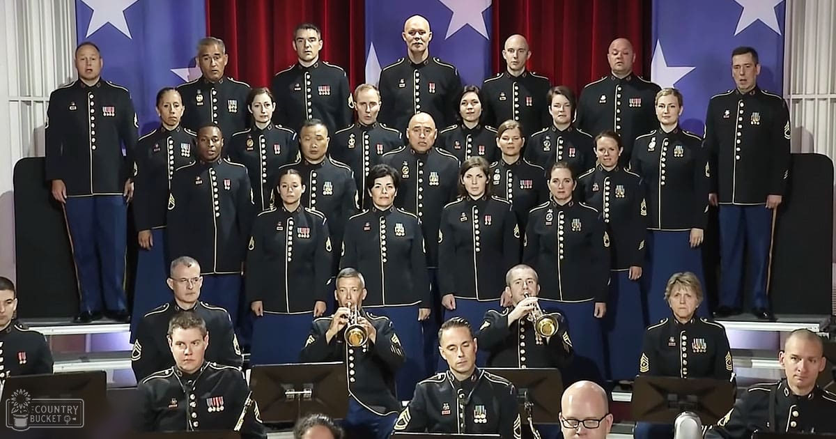 An adult male singer in a military choir performing indoors with dark blue uniforms and gold trim, set against red curtains and blue flags with white stars.