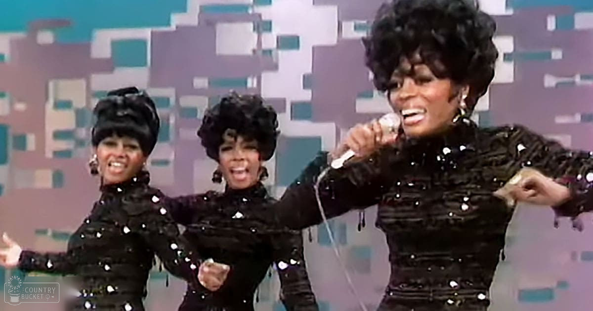 An adult female performer wearing a black sequined, fitted velvet dress with long sleeves stands on stage in front of three backup dancers dressed similarly. Bright stage lighting and colorful geometric background suggest a live performance setting.