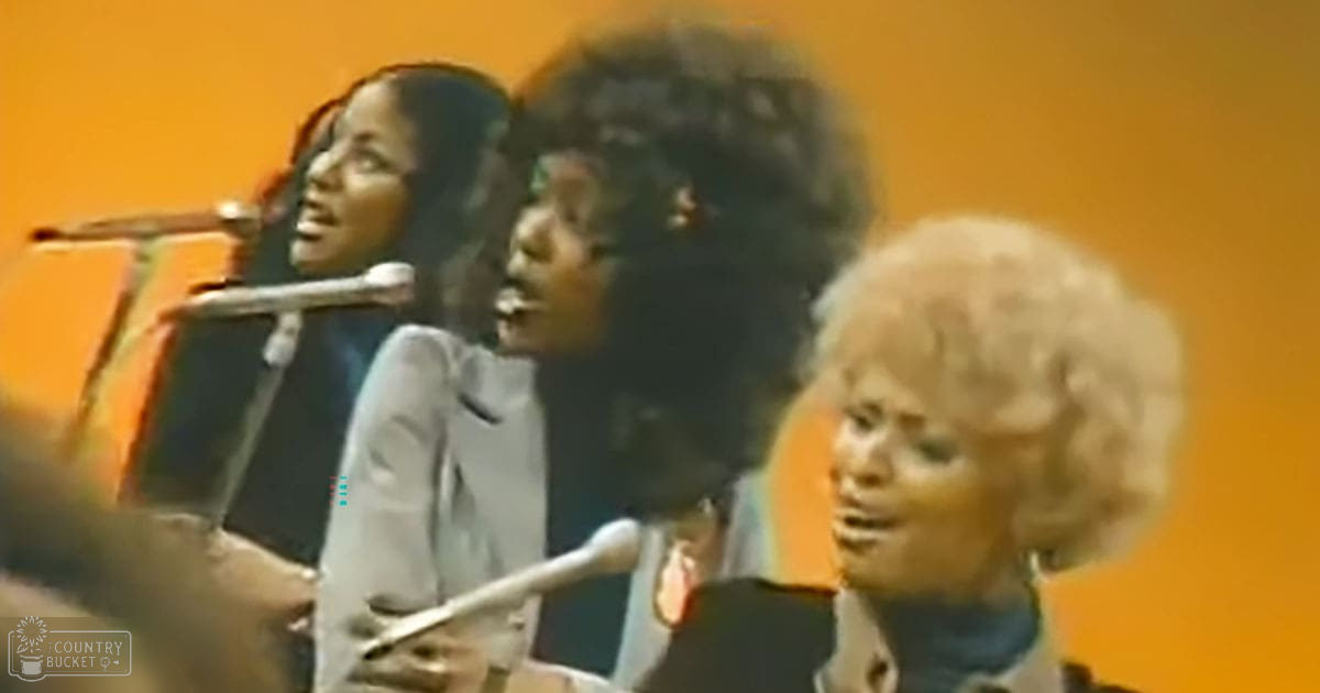 Three young adult female singers performing in a studio setting with microphones and an orange backdrop, mid-20th century style clothing.