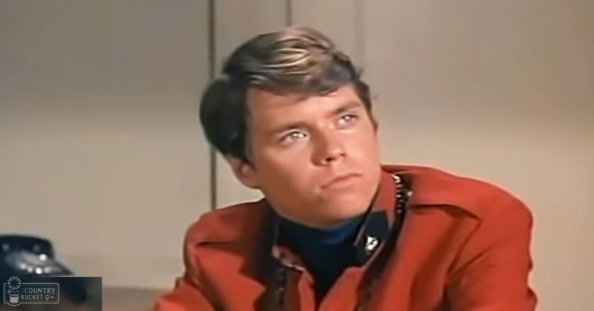 A young adult male wearing a bright red, mid-20th-century style jacket indoors with a black telephone visible in the background.