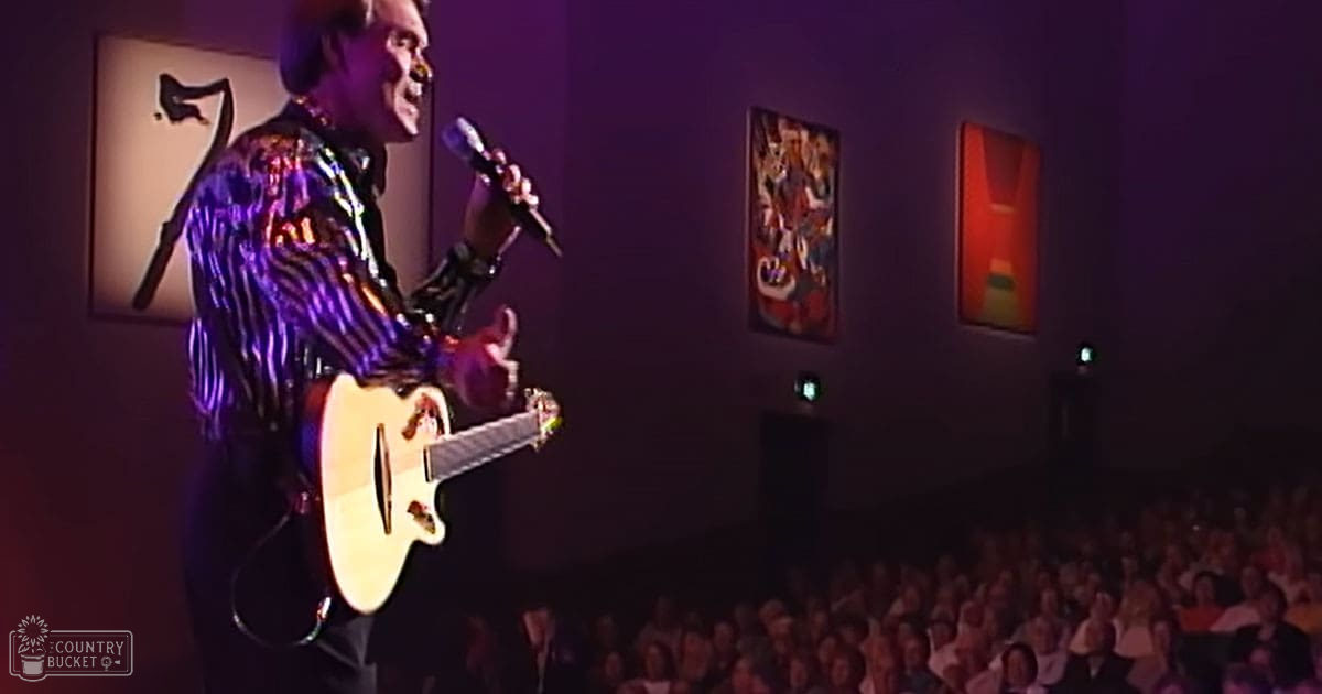 Adult male singer wearing a shiny sequined jacket with vertical purple and black stripes, performing on stage in an indoor concert hall or theater setting.