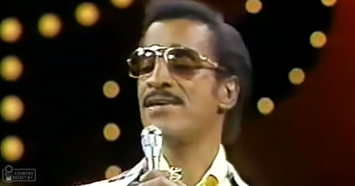 An adult male singer wearing a patterned, high-collared, long-sleeved yellow and black shirt with silk or satin fabric, holding a silver microphone in an indoor venue with warm-toned stage lighting.