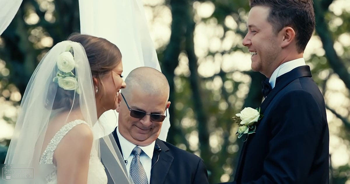 A bride in a white bridal gown with an embellished neckline stands next to her father, wearing dark sunglasses and a patterned tie, while the groom smiles in his black tuxedo. The setting is outdoors, likely in a garden or park.