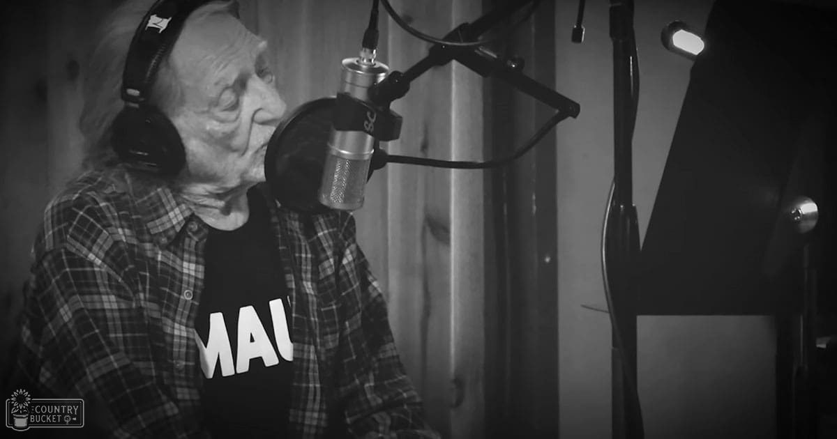 An older adult male wearing a plaid shirt over a dark t-shirt with 'MAD' printed on it, seated in a recording studio equipped with microphones and soundproofing panels.