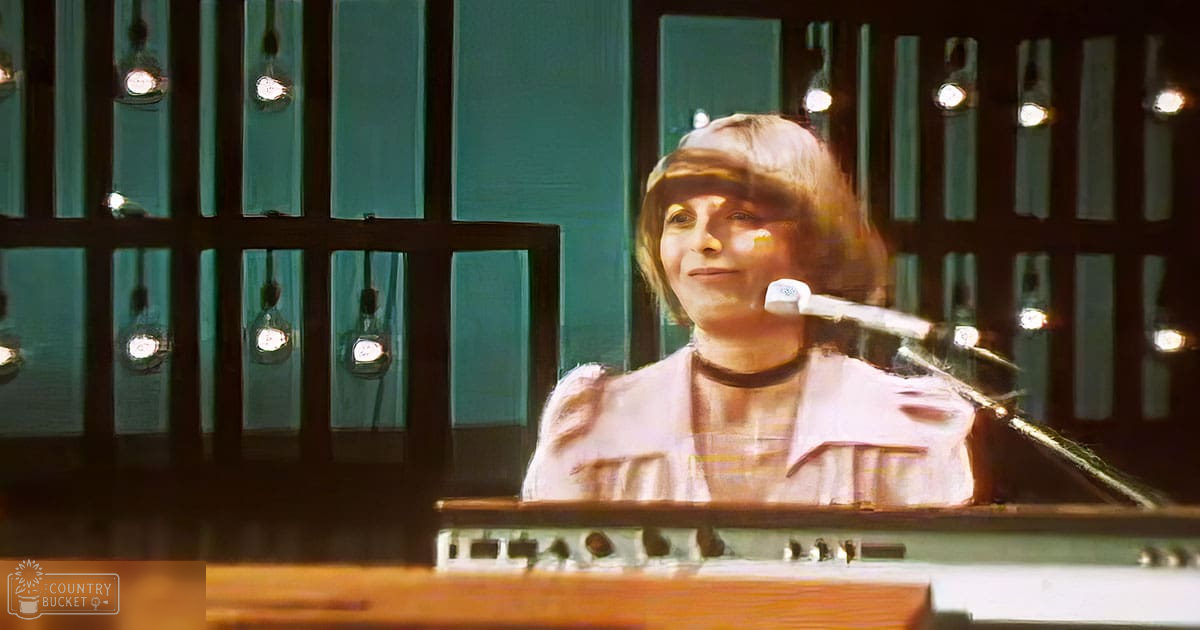 Young adult female wearing a fitted, light beige blouse with puffed sleeves and high neckline, accessorized with a choker-style necklace, seated indoors in what appears to be a recording studio setting.