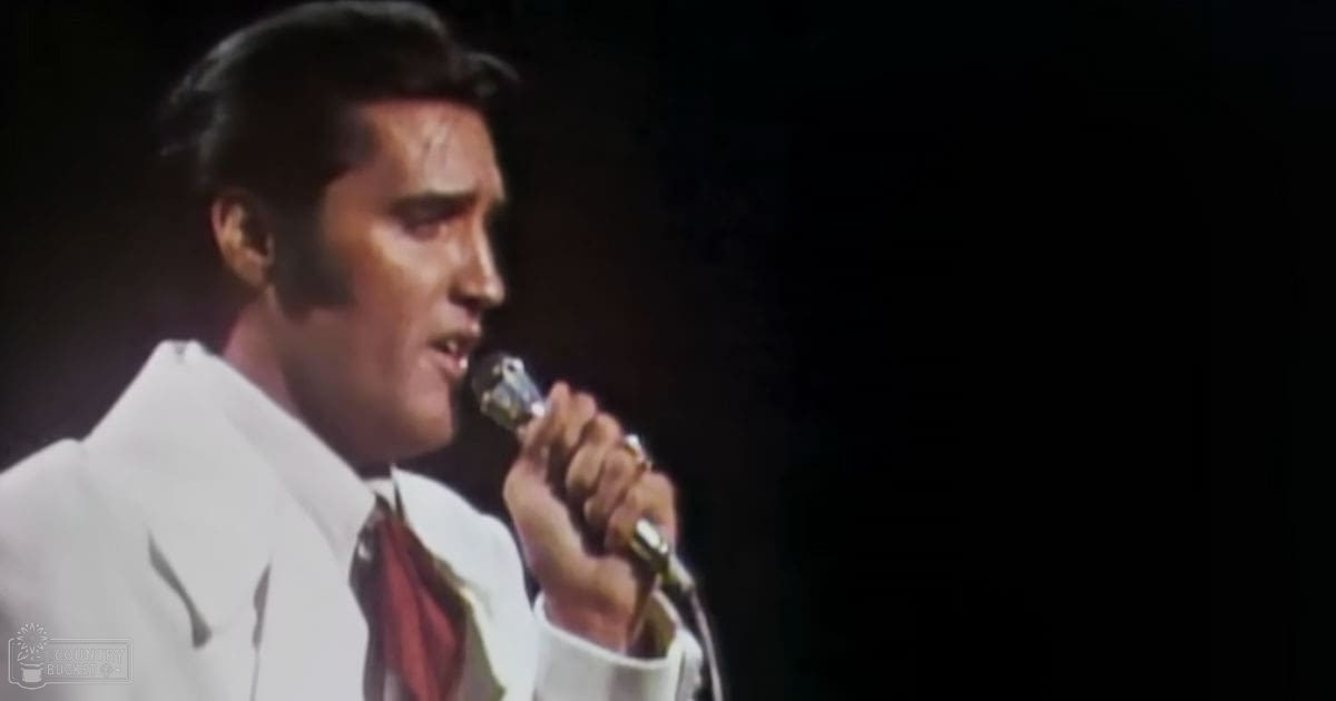 An adult male performer in a well-tailored white suit with a red tie, holding a microphone on stage.