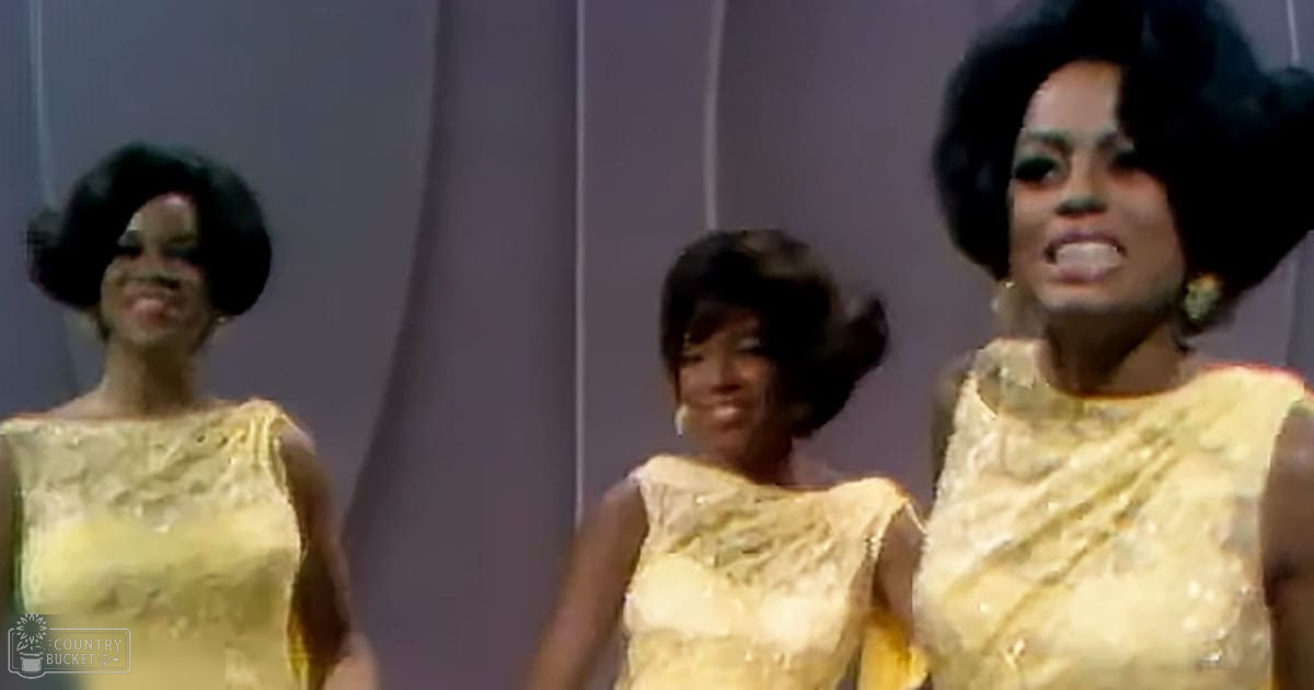 Three young adult female performers dressed in matching yellow sleeveless dresses with textured patterns, standing on a stage-like setting with plain light-colored panels.