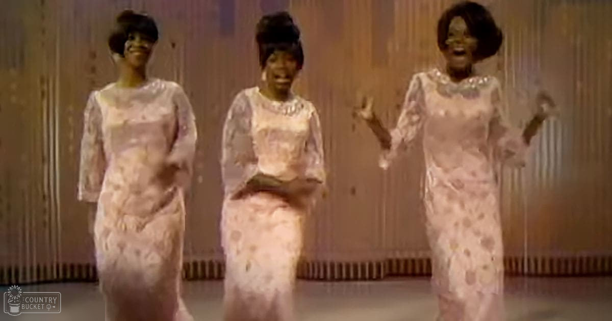 A group performance scene indoors, with adult females in long-sleeved dresses with floral lace patterns against a backdrop of curtains or drapes.