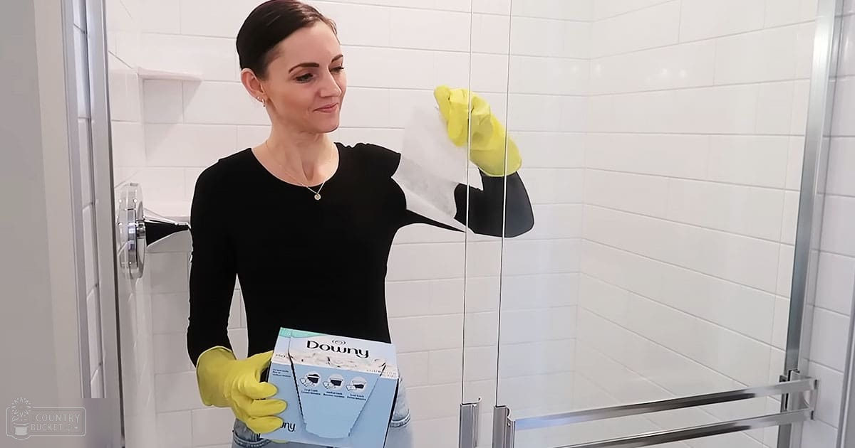 A middle-aged adult female in a bathroom, wearing a black long-sleeved top and holding a box of Downy fabric softener.