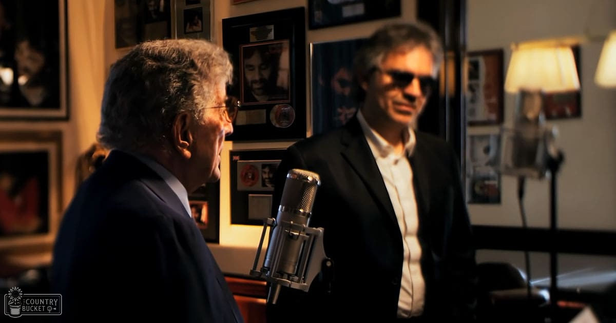 Two adult males in a recording studio setting with framed pictures on the walls, warm indoor lighting at evening or night time.