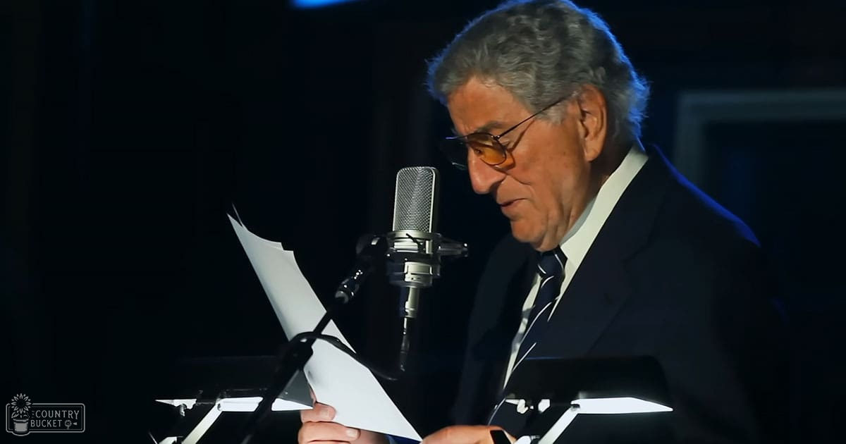 An older adult male in a formal setting, possibly on stage or at an event, wearing a dark suit jacket, white shirt, and patterned tie; holding papers with text for reading aloud.