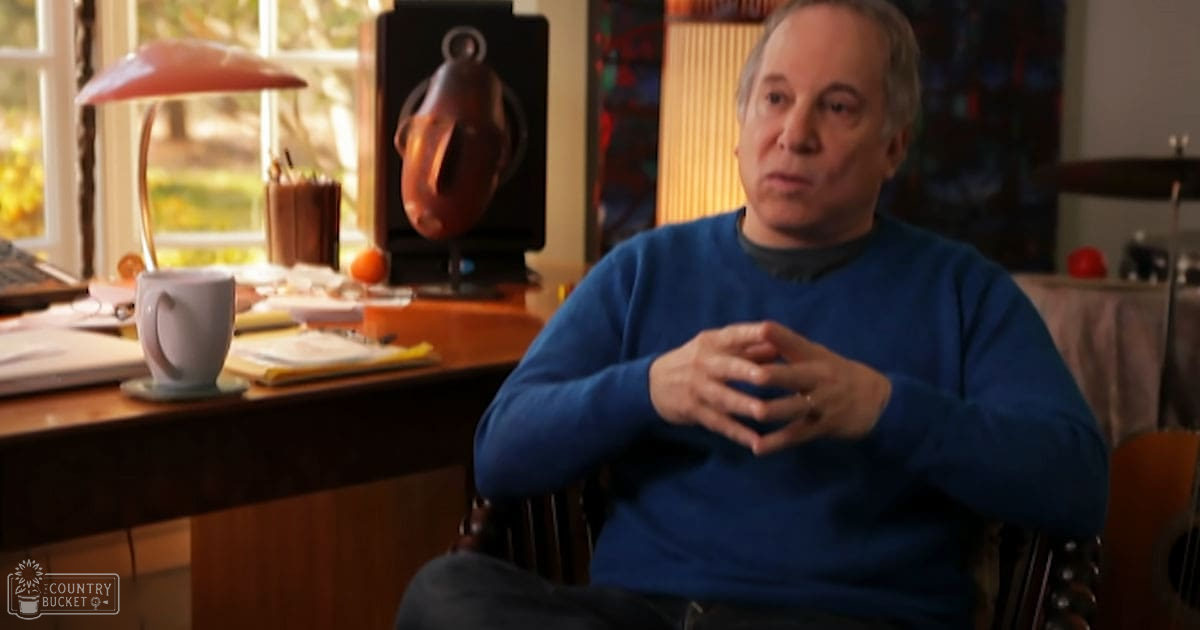 An older adult male in a home office setting with a wooden desk and various items including a red lamp, white mug on saucer, papers, notebooks, an orange fruit, black speaker, and headphones. The scene is indoors with natural light coming through the window.