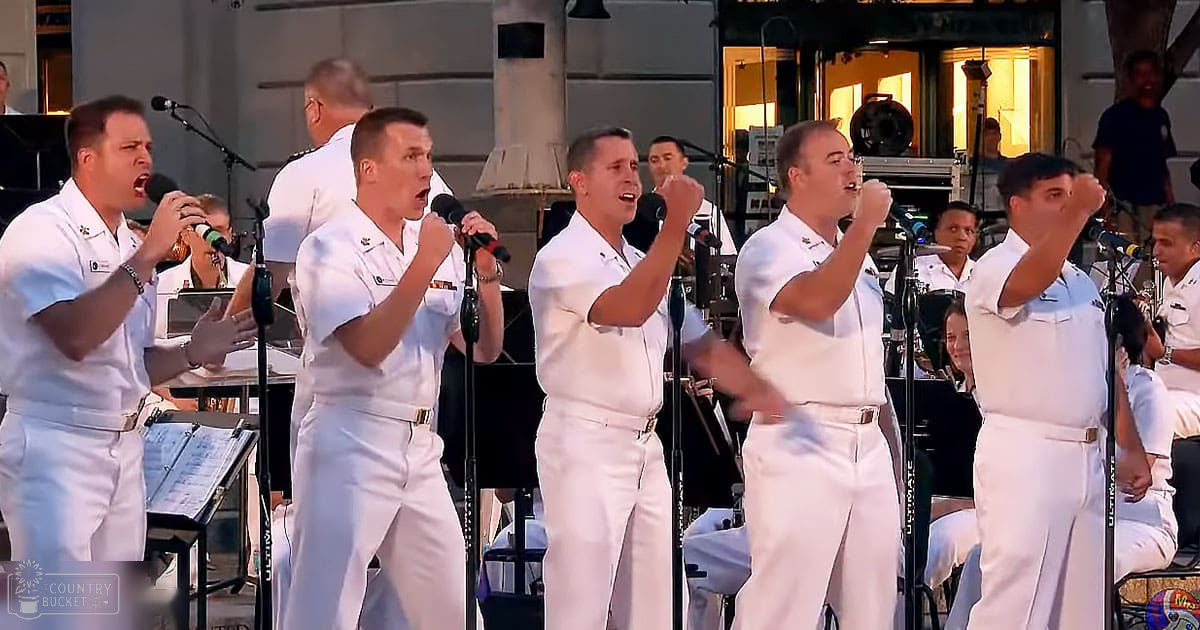 Group of male adult singers performing in an outdoor concert setting under daylight, surrounded by music stands, microphones, and stage equipment.