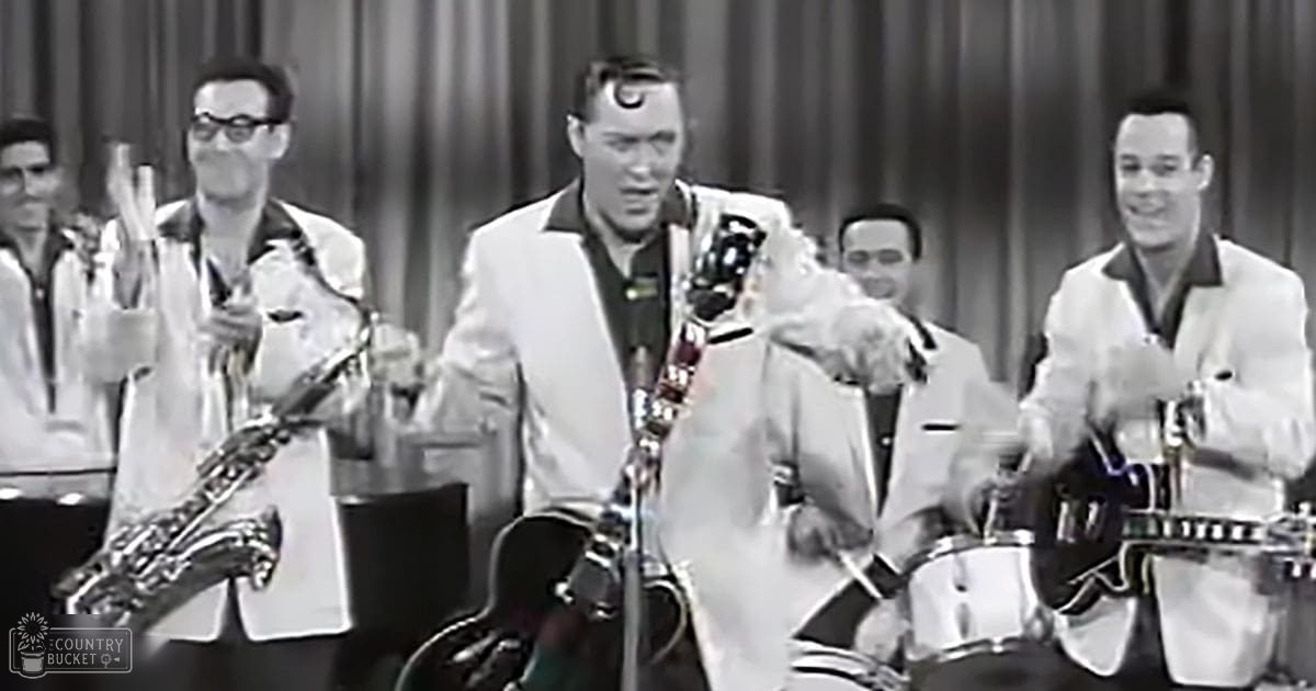 A young adult male musician in a white suit jacket performs on stage with other musicians, including a saxophonist and drummer, under stage lighting in an indoor performance venue.