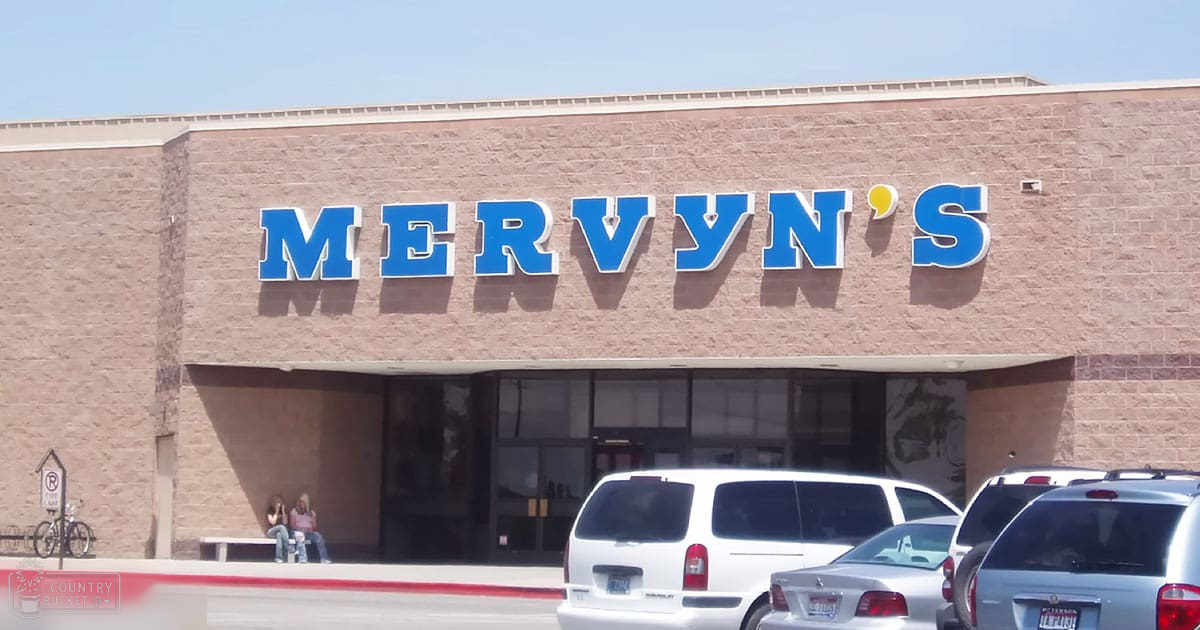 Two young adult females seated on a bench in front of 'Mervyn's', an outdoor commercial area with clear skies, likely spring or summer.