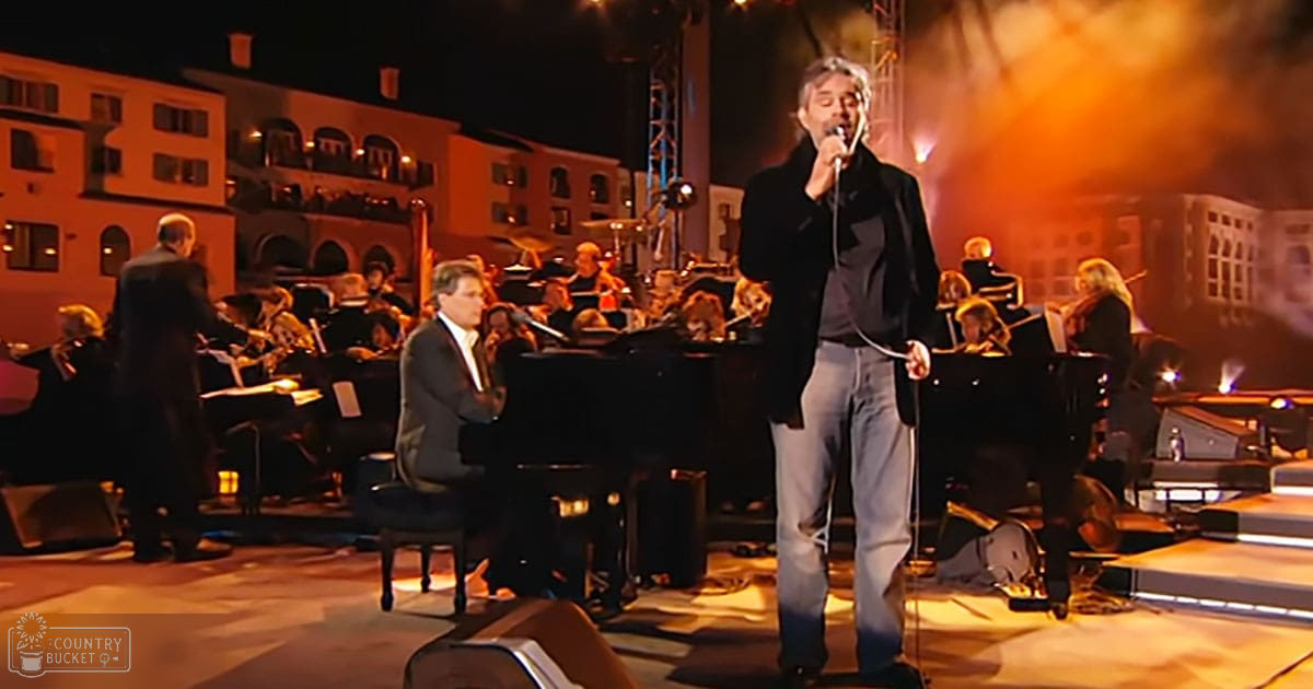 A male singer performing on an outdoor concert stage with musicians, under artificial lighting at night or evening hours.