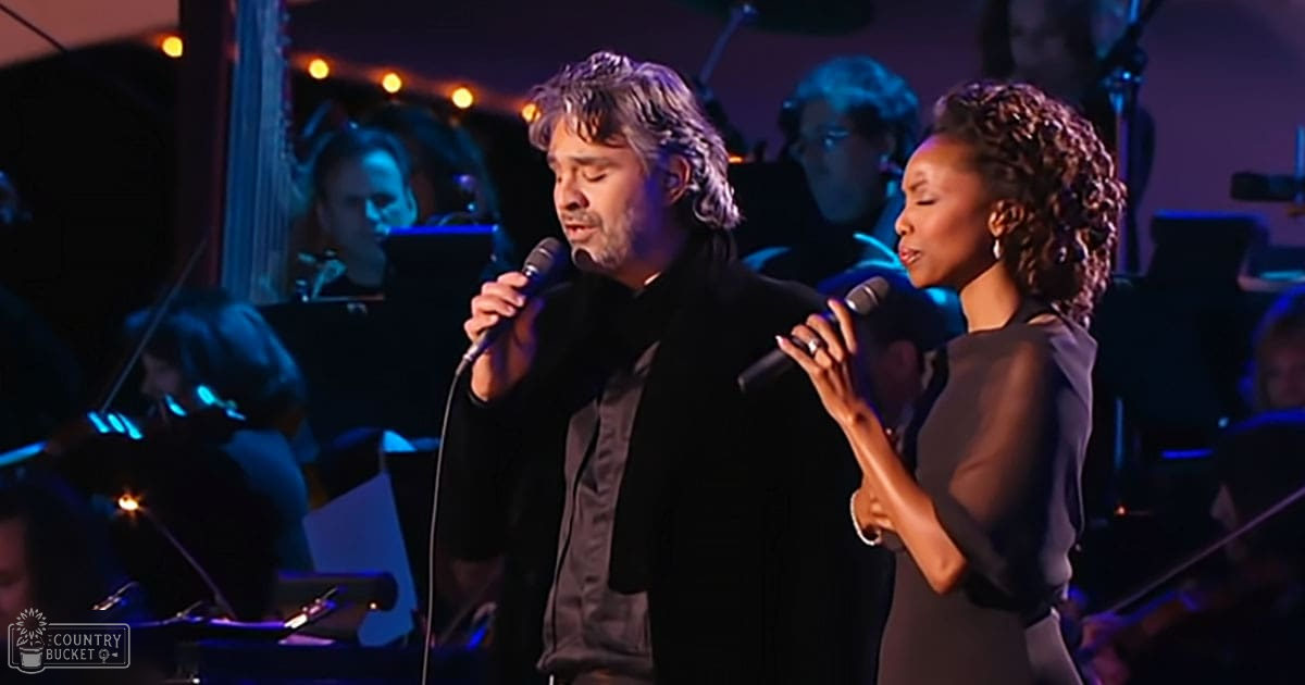 An adult male singer with shoulder-length hair, wearing a dark suit jacket over a lighter shirt, accompanied by an adult female and other musicians on stage in an indoor concert hall setting; muted lighting focuses on the performers against a backdrop of stringed instruments and percussionists.