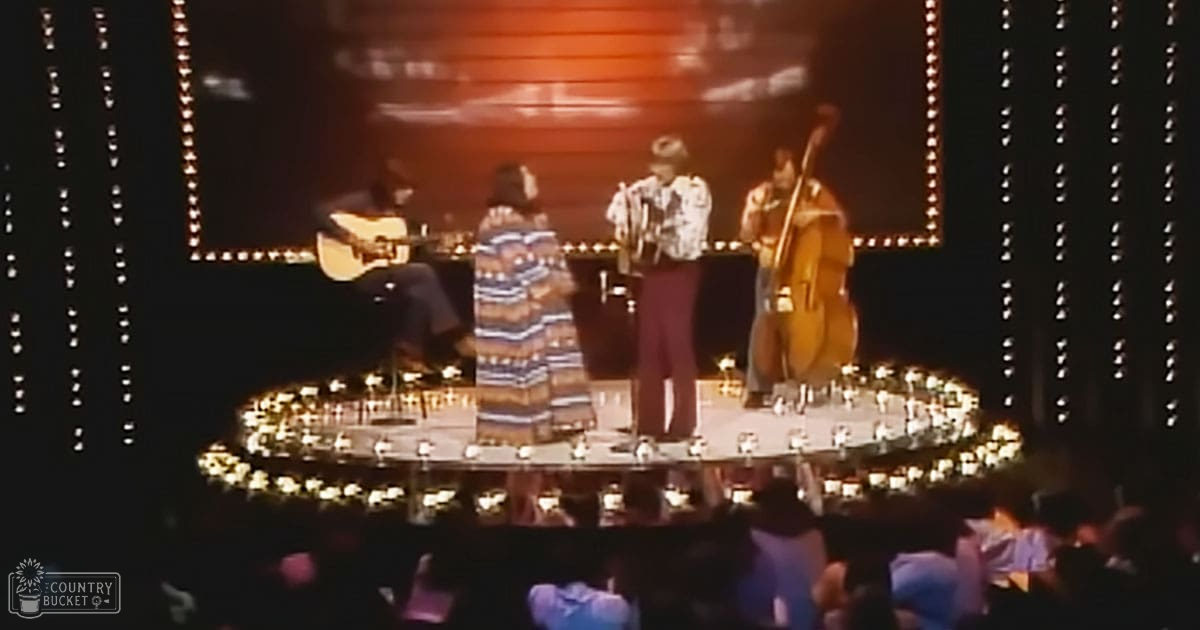 A musical performance with three adult performers on a dimly lit stage under warm lighting, wearing patterned shirts and dark pants in an indoor venue at night.