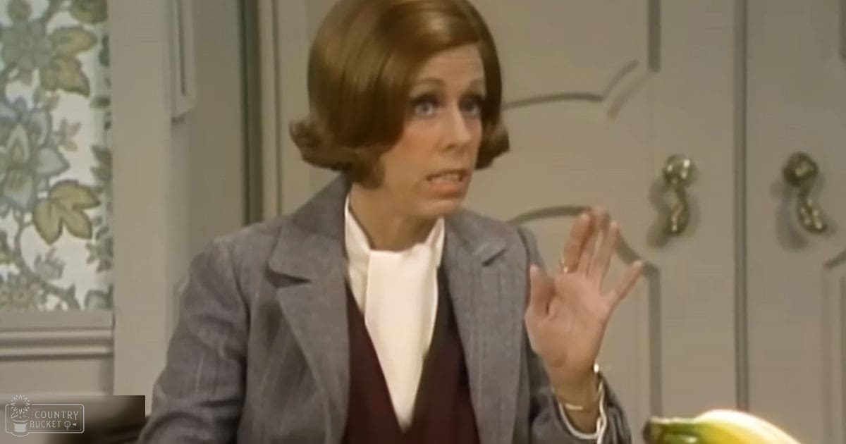 A middle-aged woman in a tailored gray blazer over a white shirt with a maroon vest, wearing accessories such as a ring, bracelet, and watch, stands indoors near light-colored cabinets and a floral-patterned curtain.