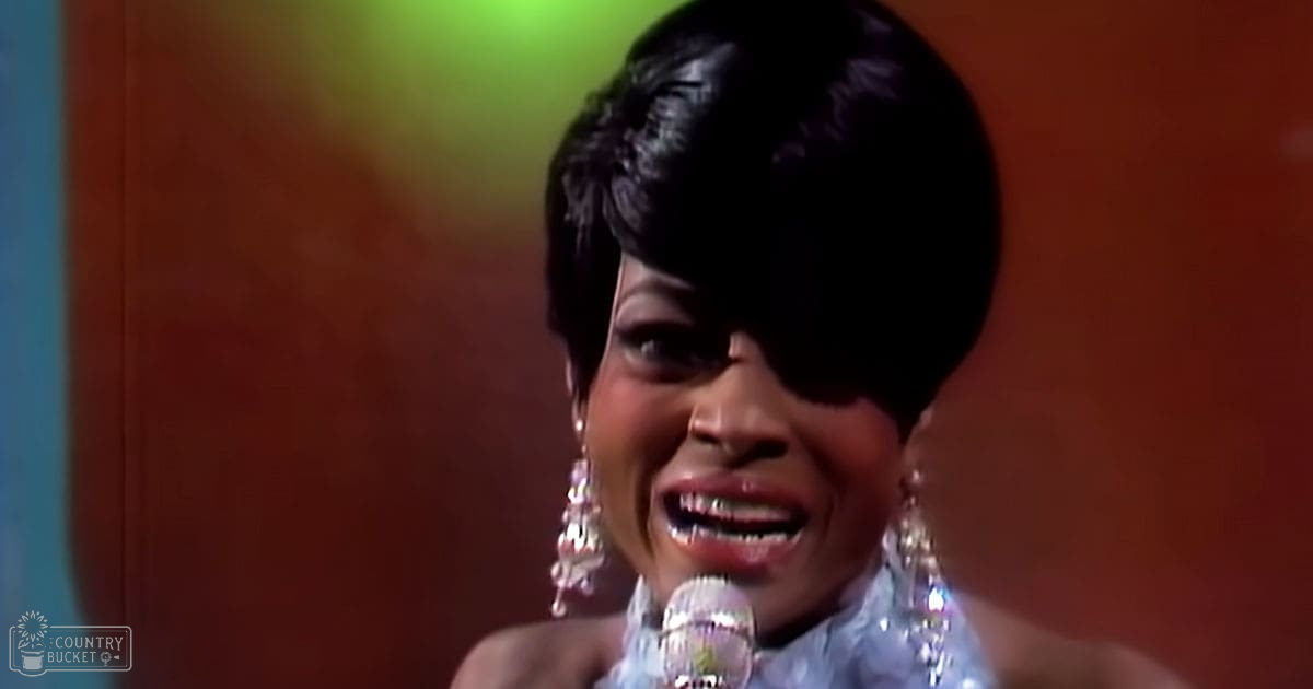 Adult female singer in a 1960s or 70s shiny garment with large dangling earrings singing into a microphone on stage under warm, soft lighting.