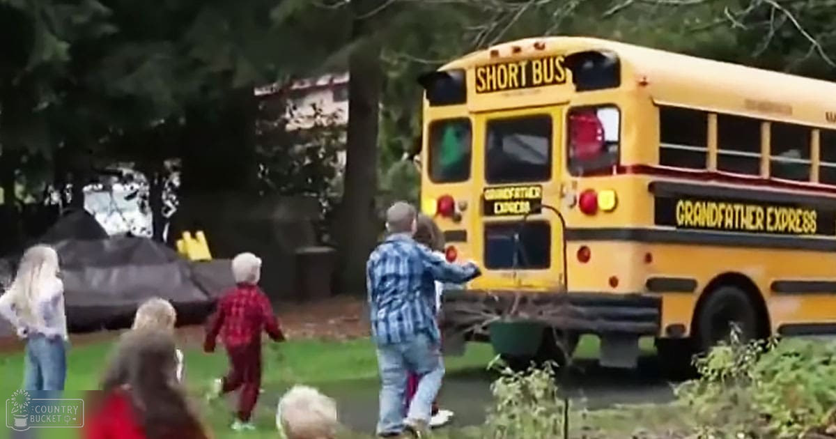 A yellow school bus labeled "Short Bus" and "Grandfather Express" is parked on a grassy area surrounded by trees. An adult male singer in a blue-and-white plaid shirt stands near the bus, facing it, while other adults and children are present in the background.