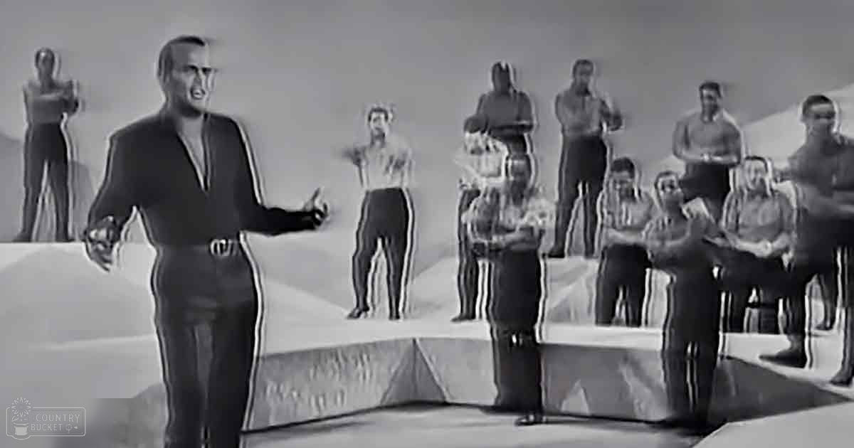 An adult male singer stands on a stage with a group of performers behind him, all dressed casually in an indoor studio setting under bright even lighting.