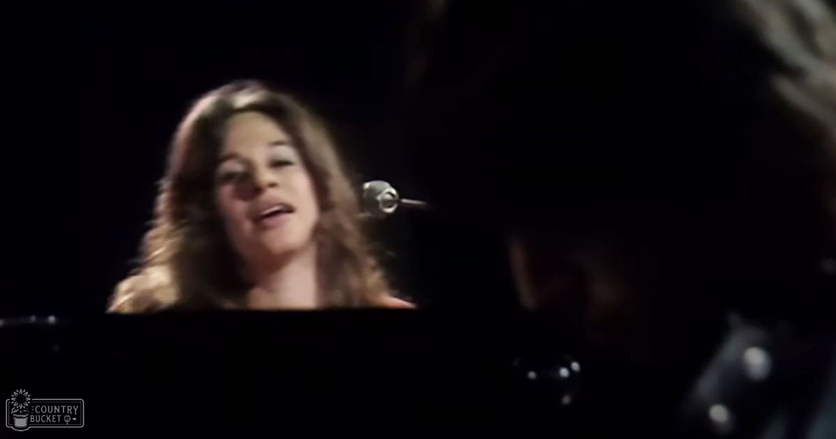 An adult female singer wearing a dark, high-necked garment at a concert setting holding a microphone.