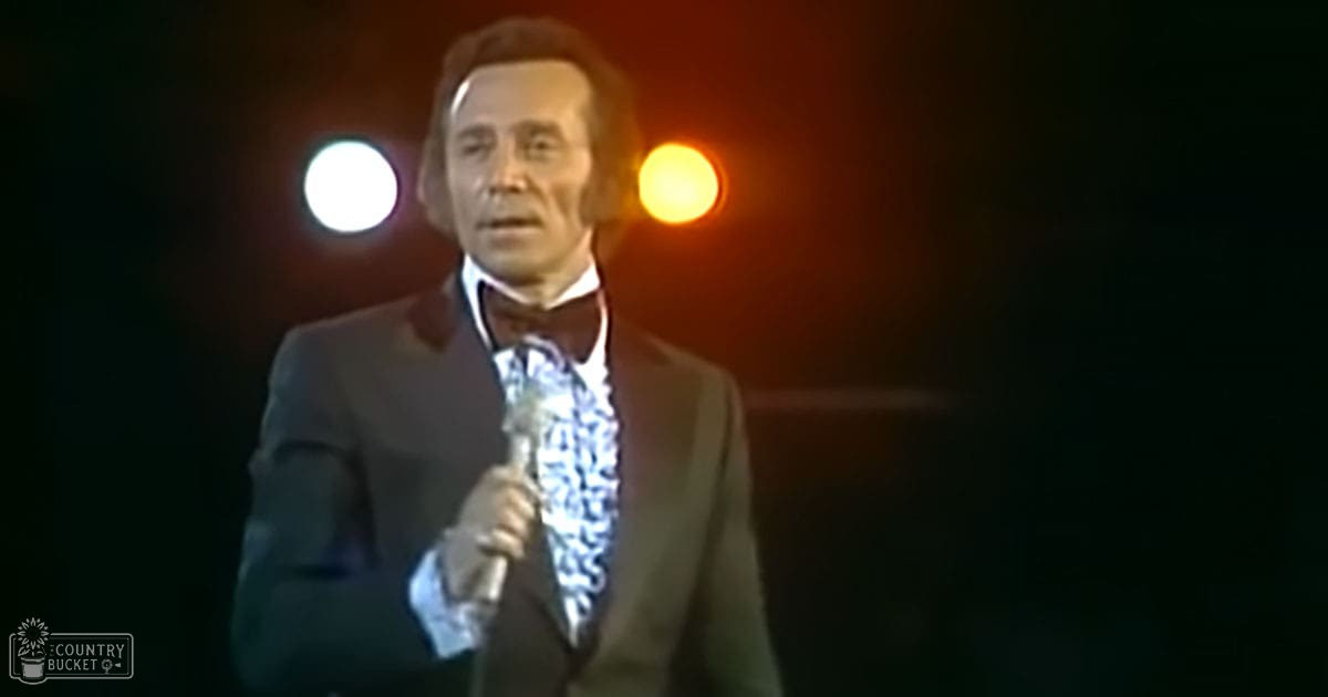 An older adult male performer wearing a dark suit jacket, white shirt, and patterned tie is on stage under dramatic lighting, with a microphone and two circular lights behind him.
