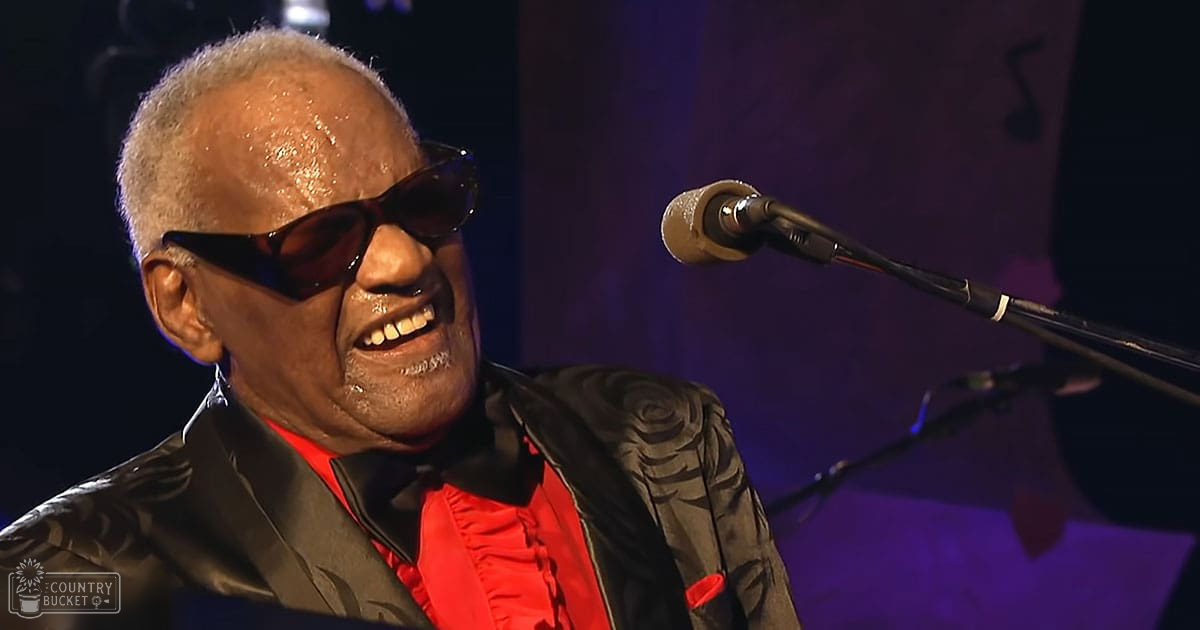 A middle-aged adult male singer wearing a black leather jacket with intricate floral patterns and red accents, performing at an indoor concert venue with dim purple stage lighting.