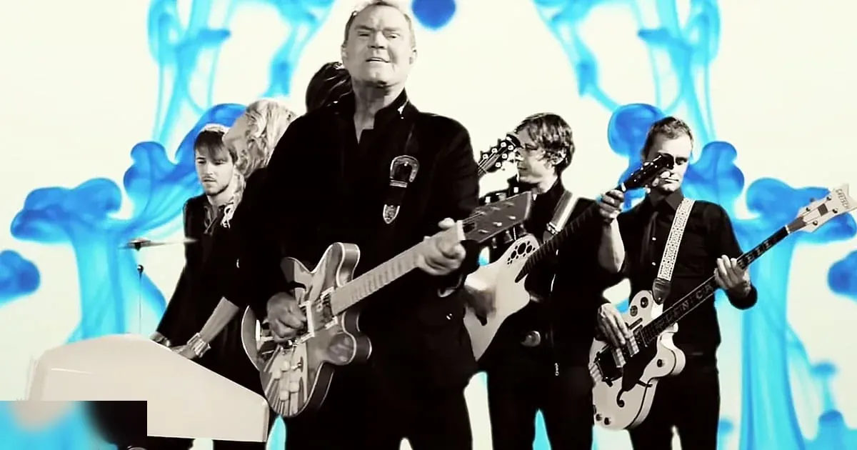 A middle-aged man playing guitar, dressed in a dark suit jacket with a distinctive patch, surrounded by musical instruments under controlled lighting.
