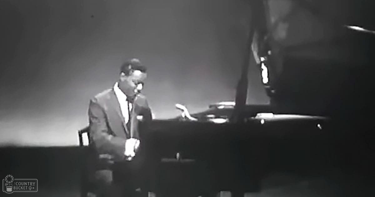 An adult male performer at a piano in a concert setting with a dark suit and formal attire, surrounded by stage equipment and dim lighting.