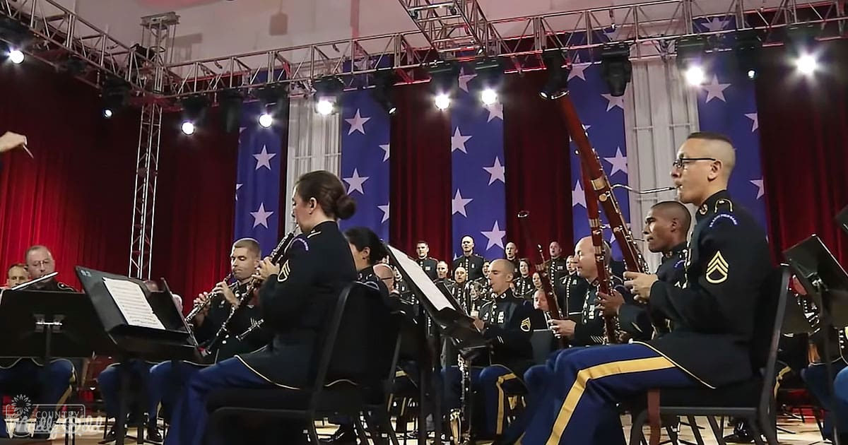 An adult male singer, wearing a dark blue military uniform with gold accents and insignia, sits among other musicians in an indoor performance setting. American flags draped over red curtains frame the stage, suggesting a formal event possibly at a U.S. military base.