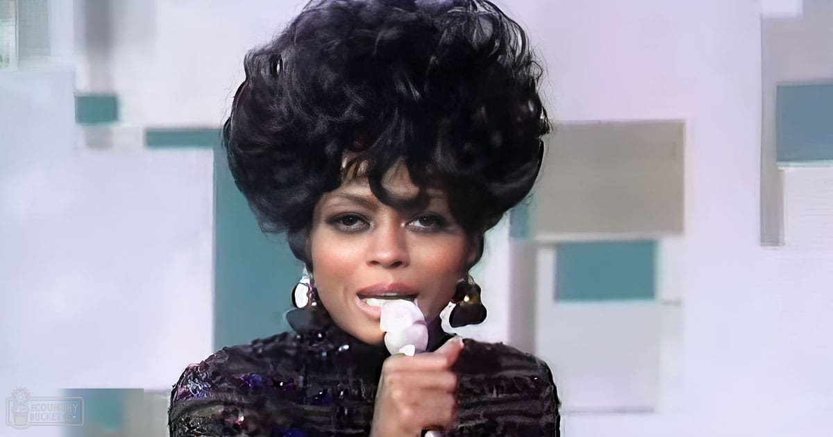 Adult female wearing a dark patterned velvet garment with long sleeves and large hoop earrings, holding a white decorative item near her mouth against a modern interior backdrop.