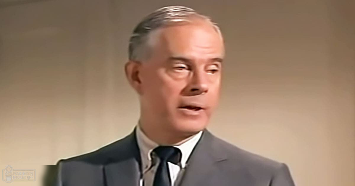 An older adult male wearing a well-fitted dark gray suit jacket with a white dress shirt and black tie in an indoor formal setting.