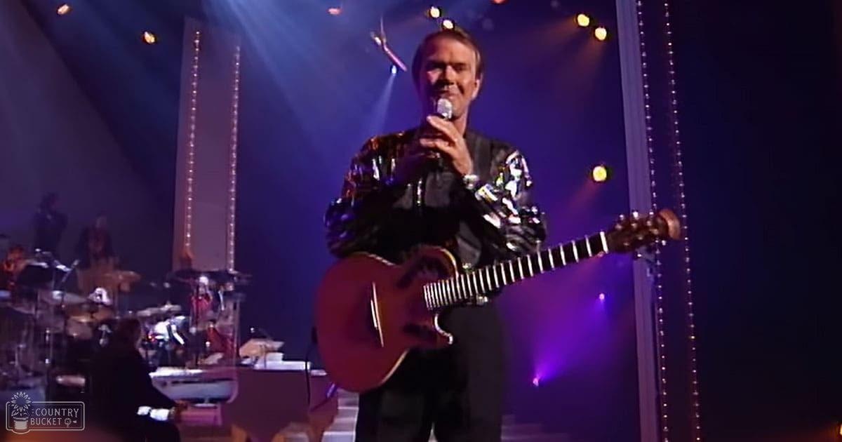 A middle-aged male singer in a shiny patterned jacket with metallic accents performs on stage at an evening concert, illuminated by dramatic purple and blue spotlights.