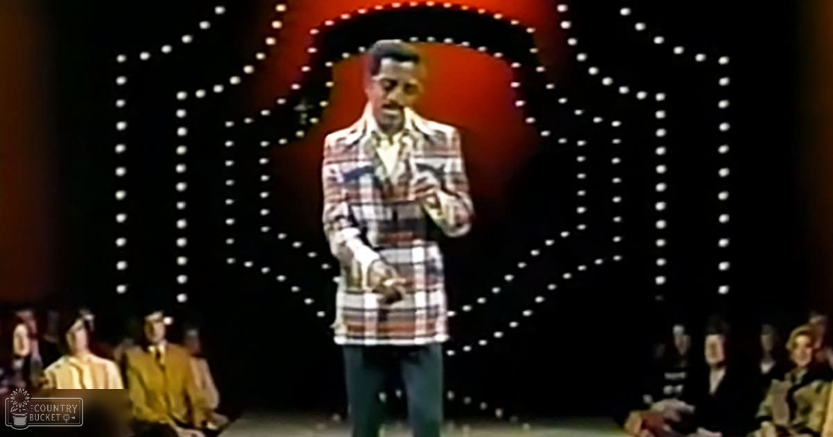 A male adult singer in a plaid jacket with white and red stripes performs on stage at an indoor comedy club or theater setting during the evening.
