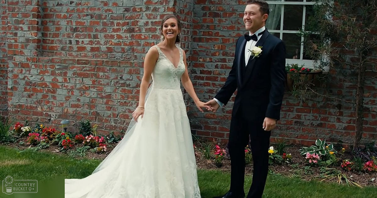 A bride and groom stand outdoors in front of a red brick wall with greenery and blooming flowers, wearing formal wedding attire against a natural backdrop.