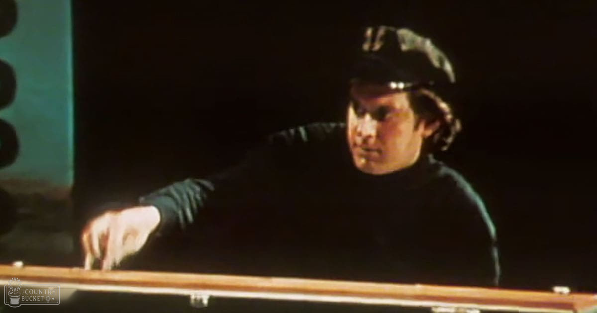 A middle-aged man wearing a dark-colored long-sleeve shirt with a high collar and possibly a black cap or hat, standing indoors under dim lighting.