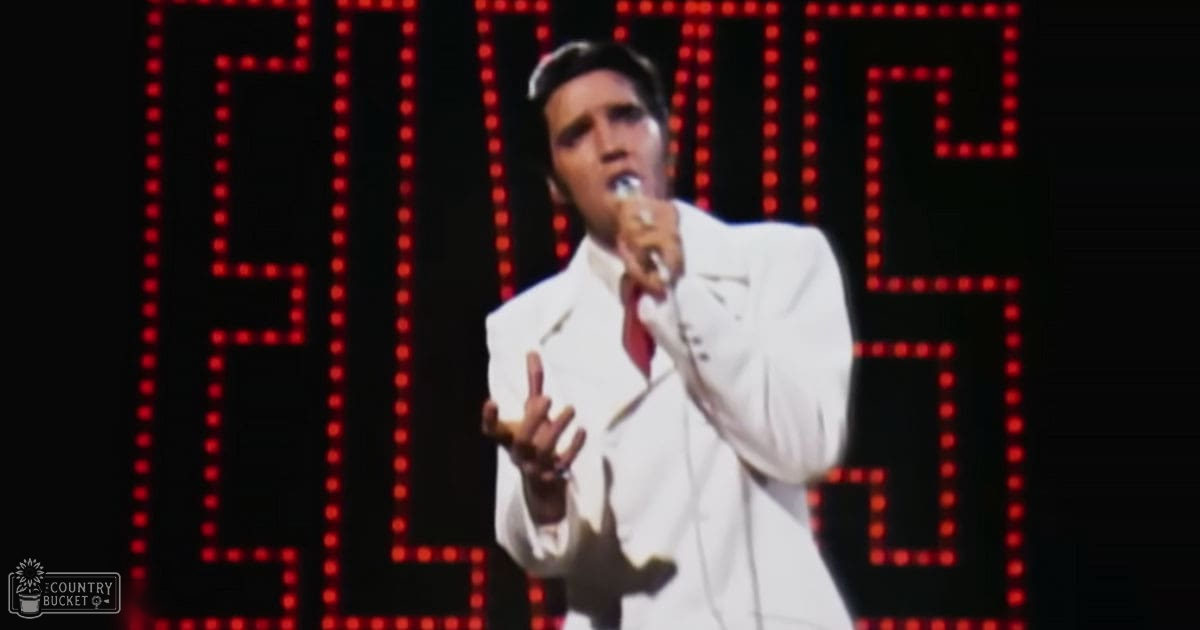 A performer in a white suit jacket, red tie, and classic fit white shirt stands on stage in front of an 'ELVIS' sign with professional lighting and large red LED letters.