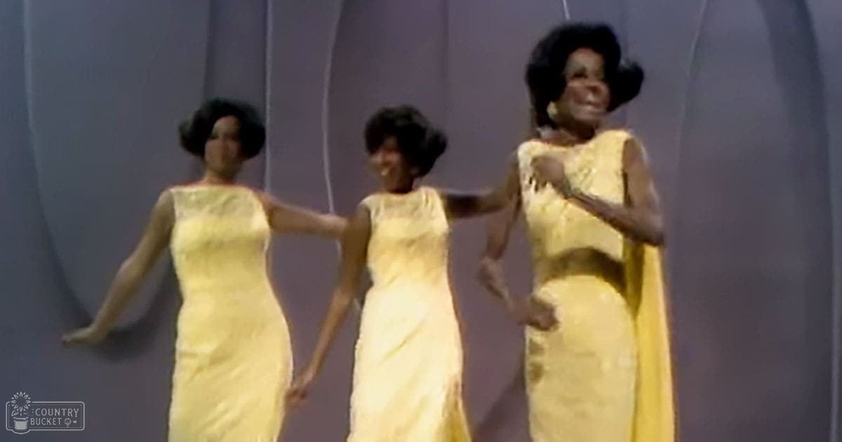 Group performance scene with adult females in sleeveless yellow dresses on a stage backdrop, mid-20th century style.