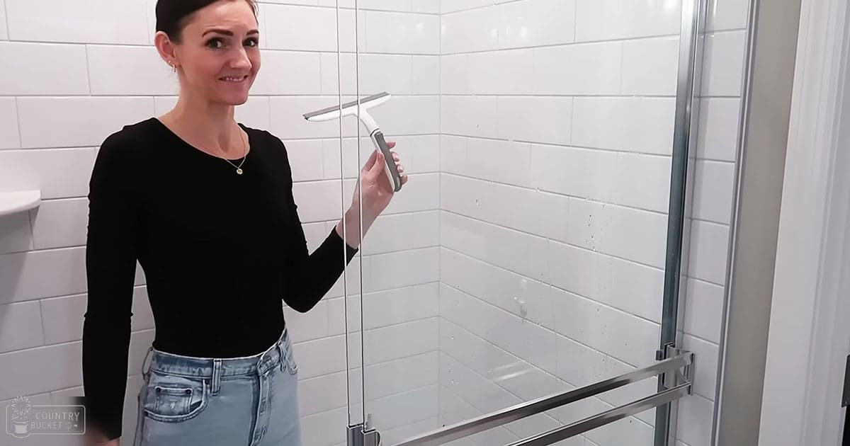 A young adult female in a bathroom wearing a long-sleeved black top with a V-neckline and a necklace, holding a squeegee near white tiled shower doors.