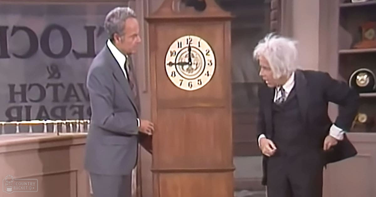 A mid-20th-century television show set with a middle-aged male in a dark gray suit and an older adult male in a black patterned tie, surrounded by furniture like a wooden grandfather clock and shelves with various objects.
