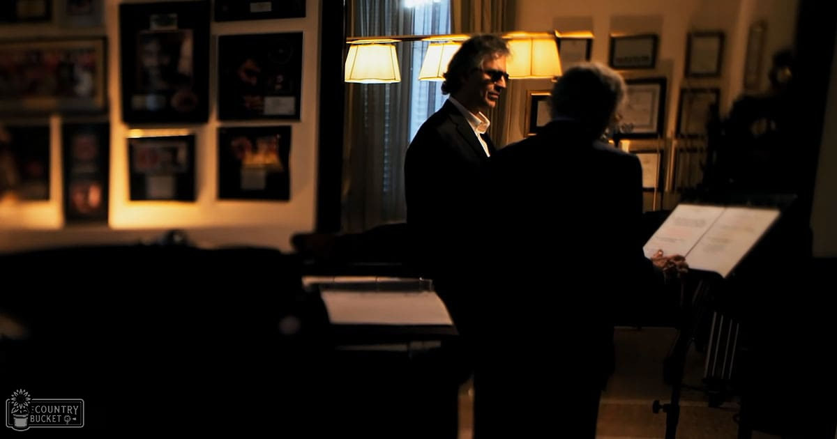 Middle-aged man in a formal office setting with older adult male seated at desk surrounded by sheet music stands and framed certificates, warm indoor lighting suggesting evening or late afternoon.