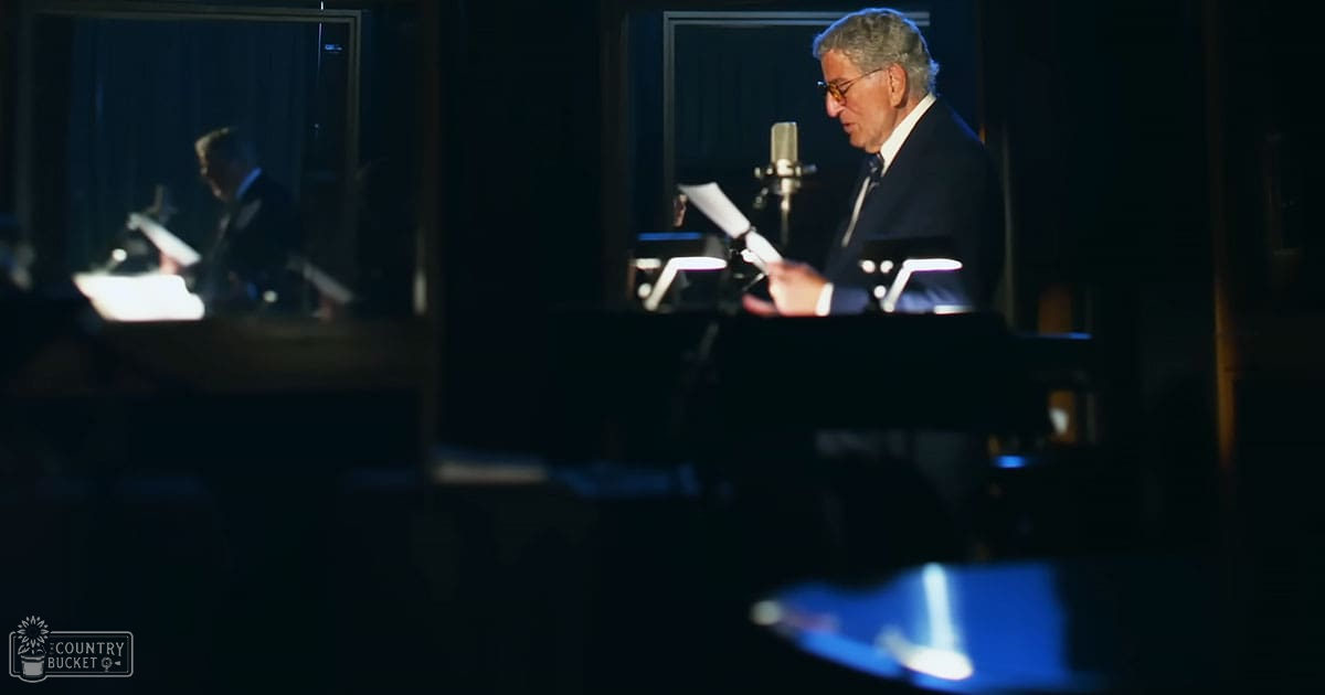 An older adult male in a dark suit jacket and white shirt, wearing glasses, stands indoors within what appears to be a recording studio with glass panels reflecting equipment and lighting.