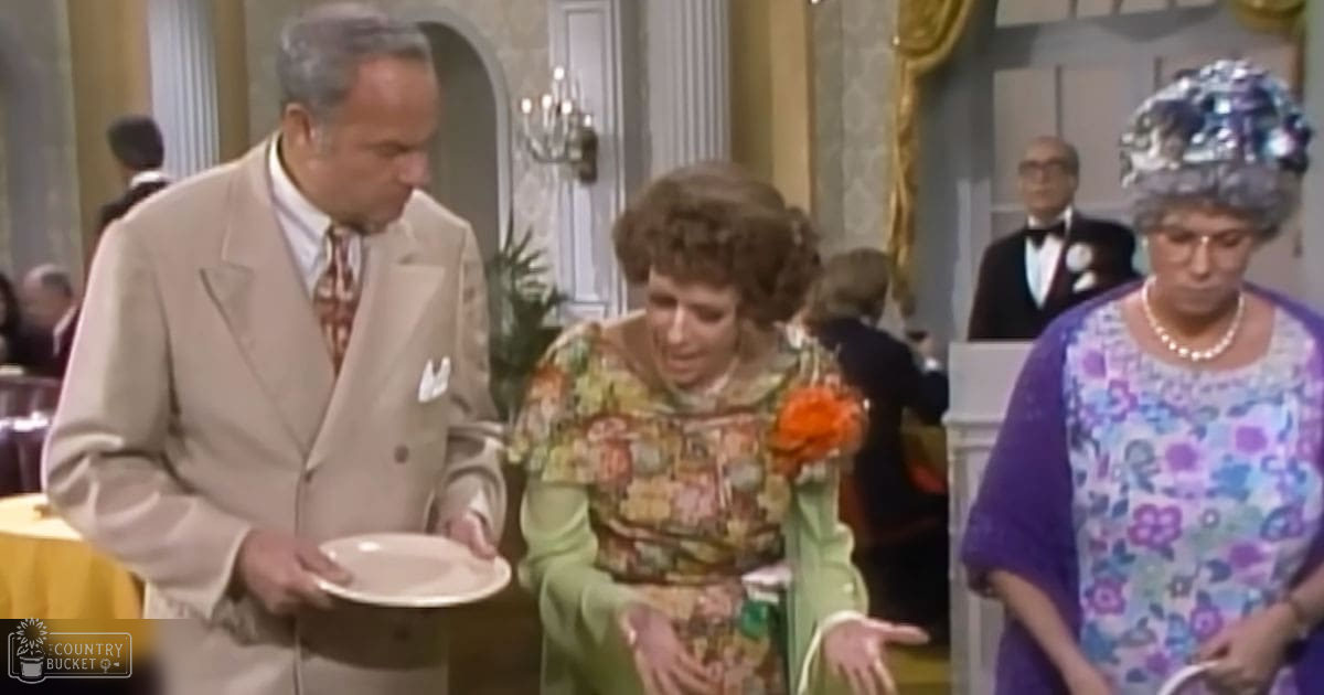 An older adult woman wearing a floral dress with green sleeves appears distressed at an indoor event where another woman in a purple floral dress and pearl necklace stands nearby, holding a plate.